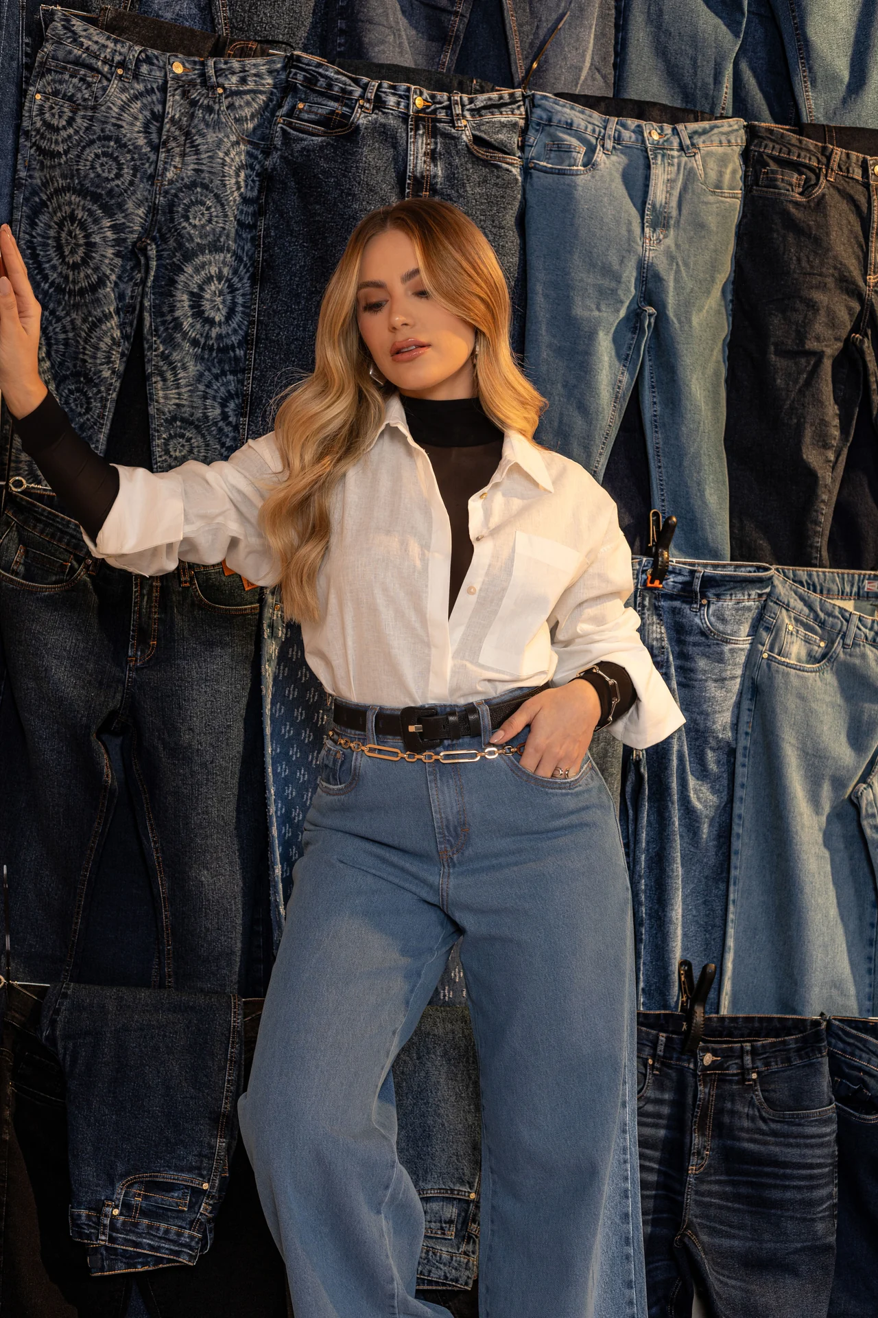 Modelo com camisa branca e calça jeans diante de um painel de jeans